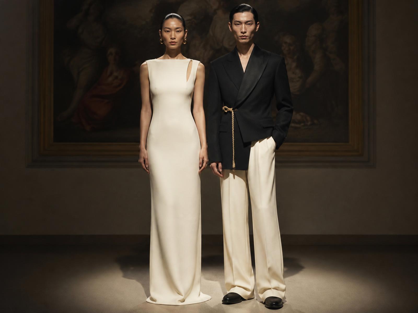 Two figures side by side in a gallery: an ivory column dress with a split boat neckline and a black asymmetric shawl blazer with a braided cord closure